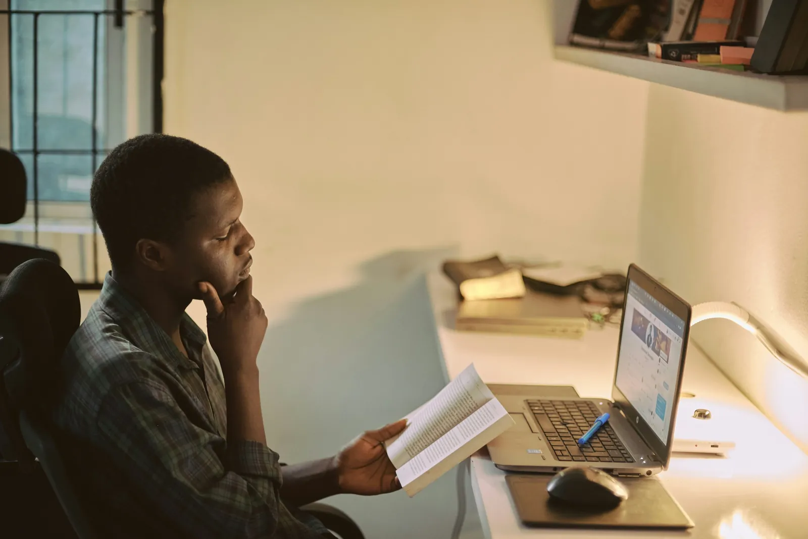 A man at a desk in warm lamp light holds an open book and rests his chin on his hand, with a laptop beside him.