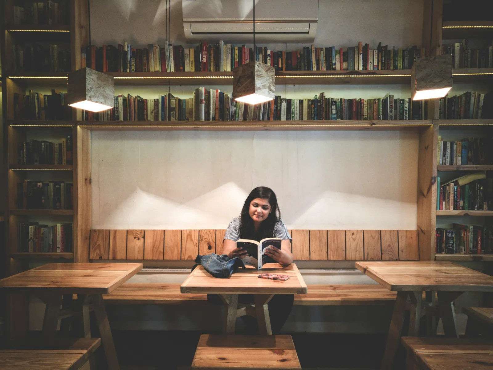 A woman reads a book at a wooden table in a library café with shelves of books behind her.