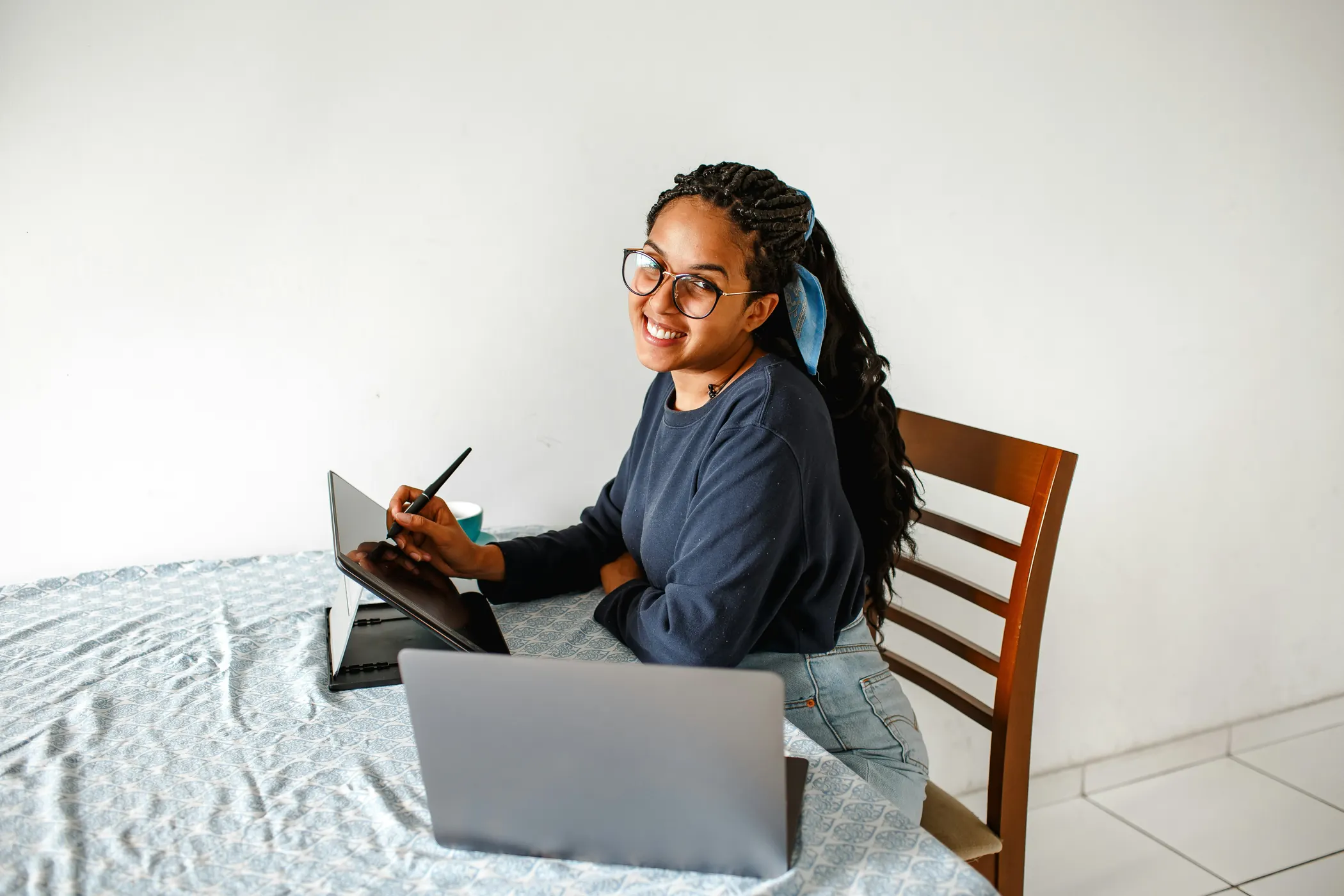 Une femme à lunettes, aux longues tresses, sourit, assise à une table avec une tablette et un ordinateur portable.